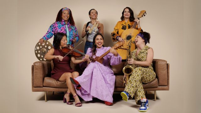 Sonia De Los Santos sits on a couch with two of her musicians. Three others stand behind the couch. All are smiling and holding musical instruments.