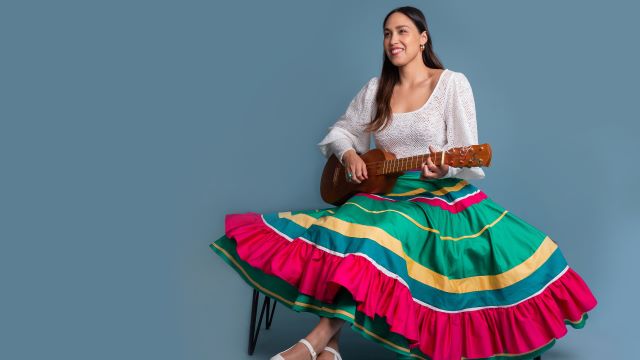 Sonia De Los Santos sits on a chair in a large, striped, colorful skirt. She is holding a guitar-like instrument and smiling.