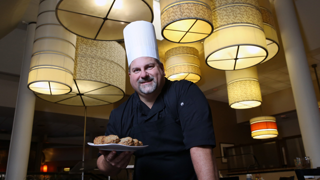 Aristo Camburako poses in a restaurant. He's wearing his chef's toque and holding a plate of cookies