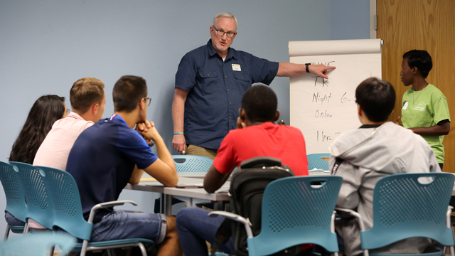 Students sitting at a table listening to a lecturer using a flipchart