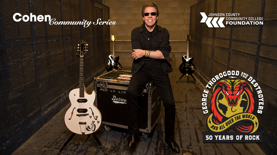 George Thorogood sits on an amp box next to his guitars while wearing sunglasses. His arms are crossed and he is smiling at the camera.