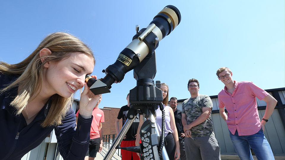 in the foreground, a student smiles while looking through the eyepiece of a telescope pointed toward the blue sky. In the background six other students stand in a line behind her and smile toward the camera.