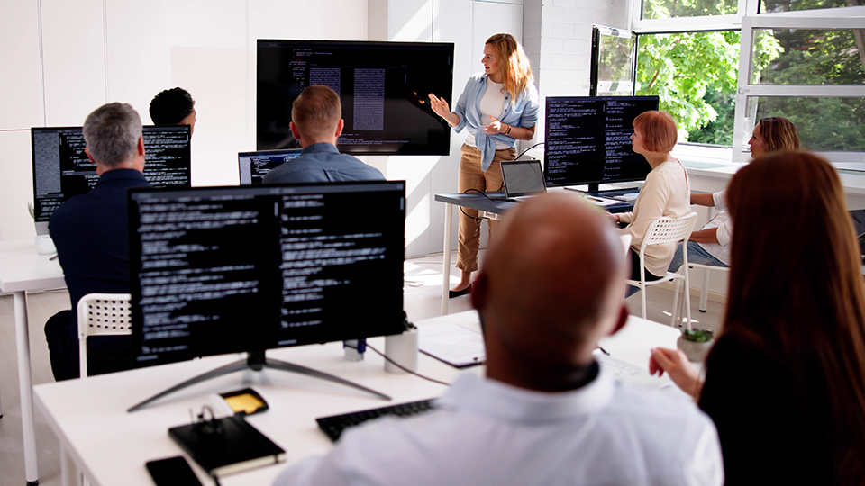 A classroom filled with students working on a computer and an instructor at the front of the room teaching with a large computer screen behind her.