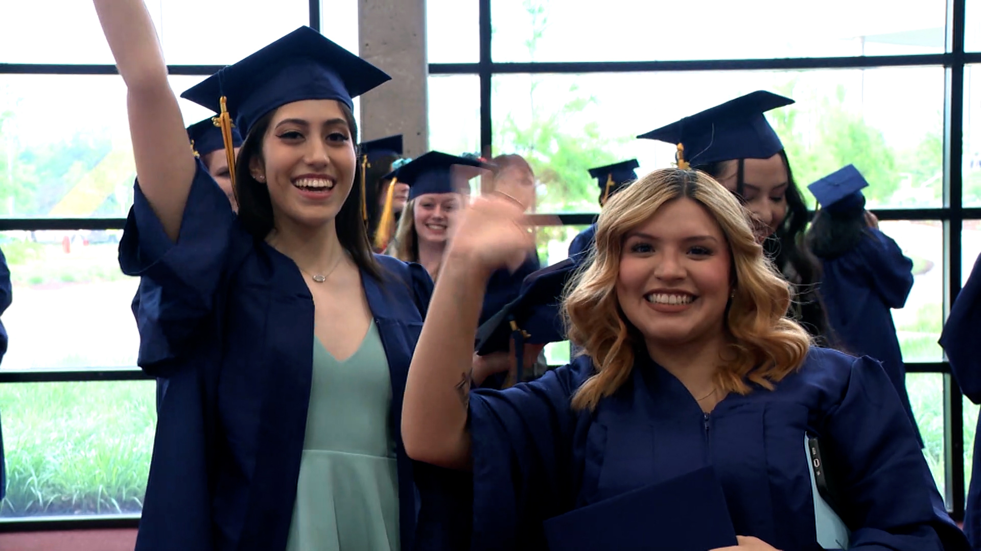 Two graduates in caps and gowns waving at the camera.