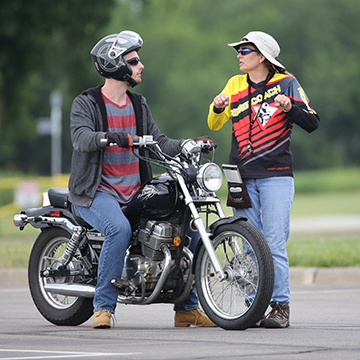 A motorcycle teacher demonstrates proper handlebar grip technique to a student who is astride his bike.