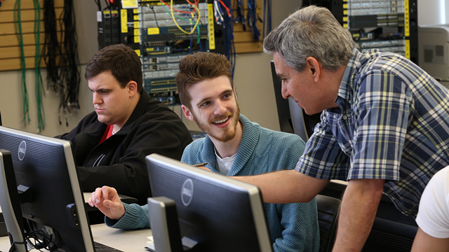 2 smiling students working on a computer as an instructor leans down to help