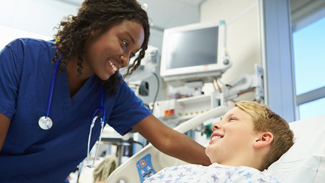 A nurse in blue scrubs smiles at a young boy in a hospital bed.