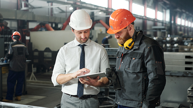 A man in a white hardhat looks at an e-tablet while a man with an orange hardhat looks over his shoulder.