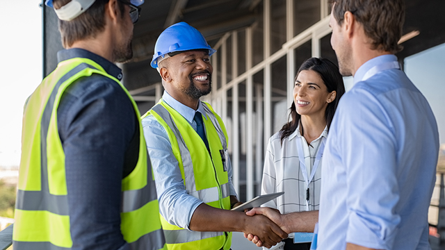 A man in a blue hard hat and safety vest shakes hands with a man in a button down shirt while a woman looks on and smiles.