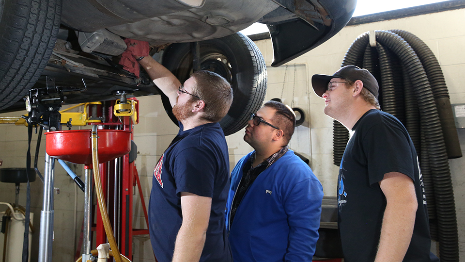 Three men look at the underside of a car that is on a lift.