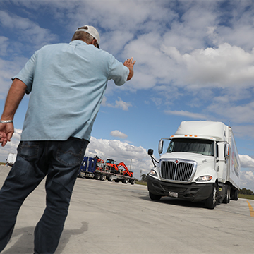 A cdl instructor stands with his back to the camera and hand raised while directing a student driving a semi truck