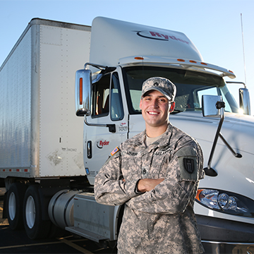 A male veteran in uniform with arms folded looks at the camera and smiles while standing in front of a semi truck