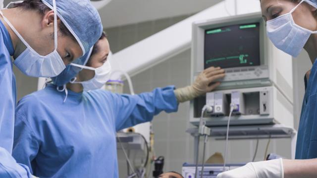 A person in a surgical mask points at a monitor in a surgical suite while another person looks on