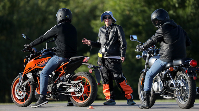 A motorcycle instructor gestures with one hand raised while talking to two students sitting on motorcycles