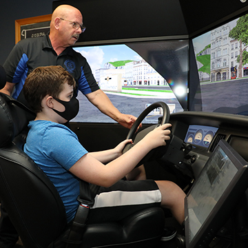 masked student sits in a driving simulator while an instructor stands behind him