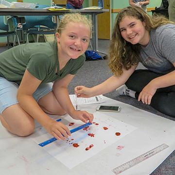 two female students sit on the floor while measuring on a large piece of white paper