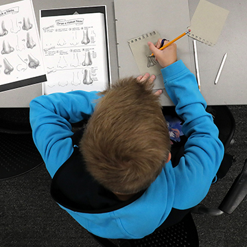 overhead shot of a boy sitting at a desk drawing human noses