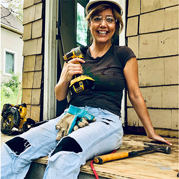 Young woman in a hard hat holding a drill sitting on a porch of a house for Habitat for Humanity