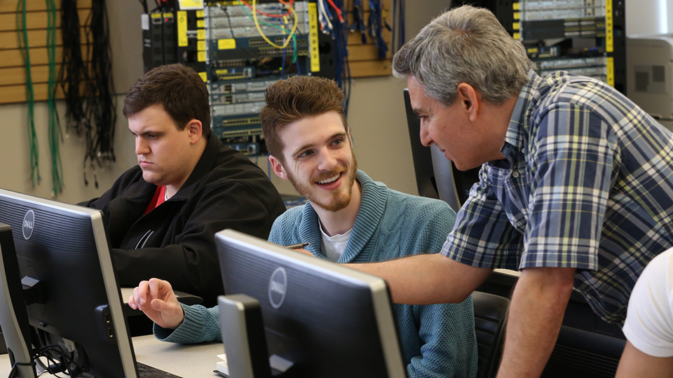 A student talks with his professor in a networking class while another student works on a project.