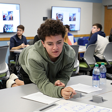 A student leans across a work table to see what another student has written.