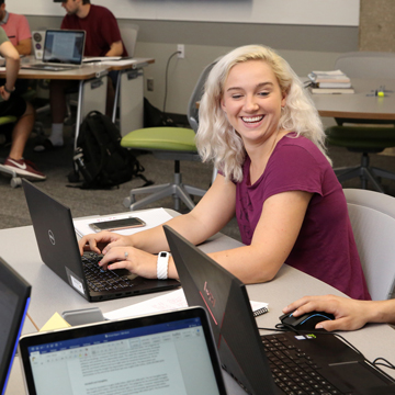 A student smiles while glancing at the computer screen of another student.