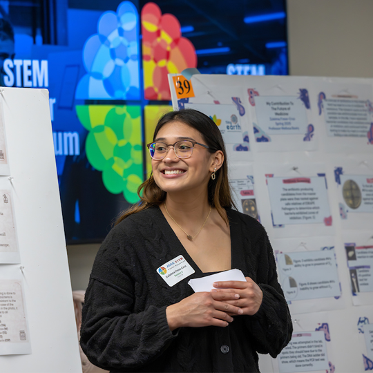 Student, smiling, standing in front of her project at the STEM poster symposium