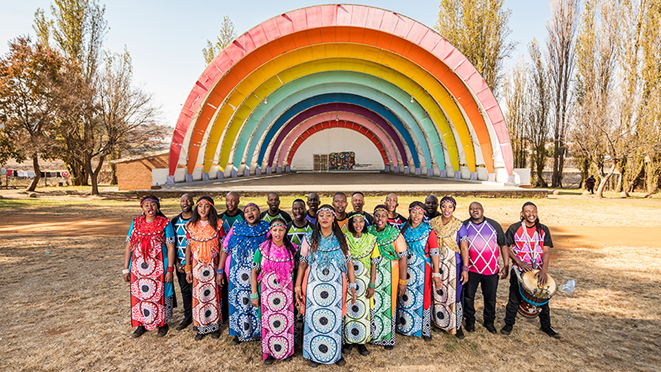 Soweto Gospel Choir members wearing brightly colored costumes and standing in front of a brightly colored outdoor stage