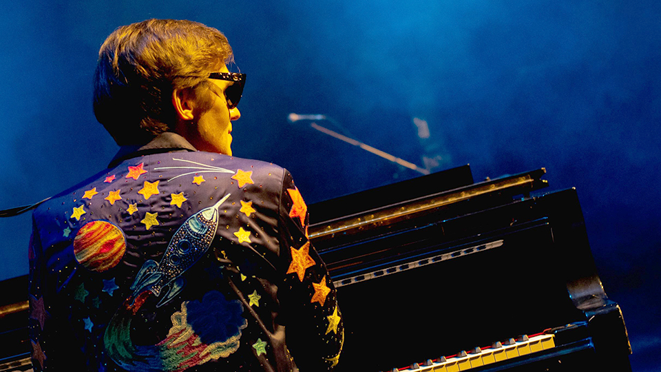 a man dressed as Elton John is seen from behind and sitting at a piano