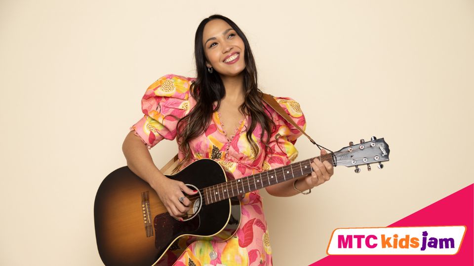 Sonia De Los Santos stands against a beige background, and smiles while holding a guitar. She is wearing a floral dress.