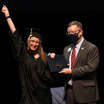 A GED graduate wearing a cap, gown and face shield receives her diploma from JCCC president Dr. Andy Bowne while smiling at the camera and raising a fist in the air