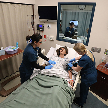 2 nursing students practice care on a simulation dummy while being observed by a nursing faculty member