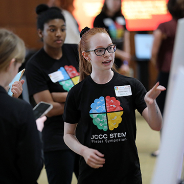 A JCCC student wearing a STEM poster symposium t-shirt points to a poster on an easel