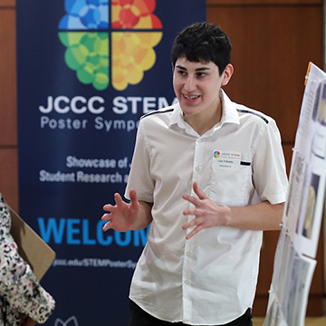 A JCCC student standing in front of a STEM poster symposium banner explains his research
