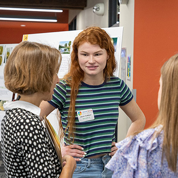a red haired student smiles at two attendees who are reviewing her poster