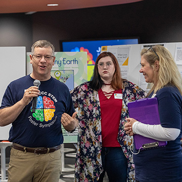 A man in a STEM Poster Symposium t-shirt holds a microphone while speaking about the event. Three smiling event organizers stand near him.