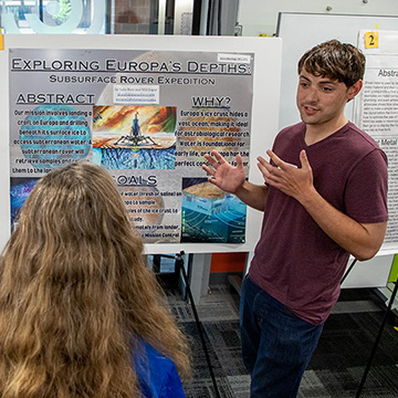 A student explains his poster titled Exploring Europa's Depths to two women who are listening intently