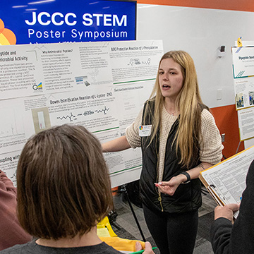 A young woman explains her research to a group of judges at the STEM Poster Symposium at JCCC