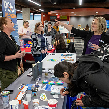 A woman points at a sign for PGAV architects while a representative of that company smiles at her. In the foreground a student is signing a piece of paper on a table.