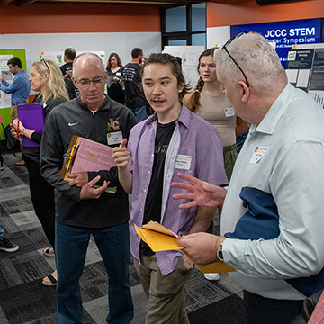 A young man explains his poster to a group of three older men at the 2024 STEM Poster Symposium