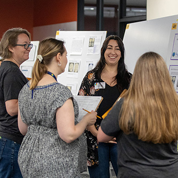 A smiling woman stands next to her poster presentation. She is surrounded by three other women who are judging her work.