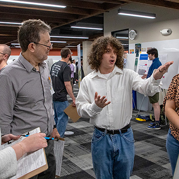 A young man gestures with his hands while explaining his poster research to a group of onlookers.