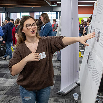 A woman points to her research poster at the 2024 STEM Poster Symposium at JCCC