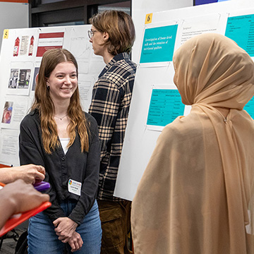 A young woman smiles politely at a group of people gathered around her poster at the STEM Poster Symposium