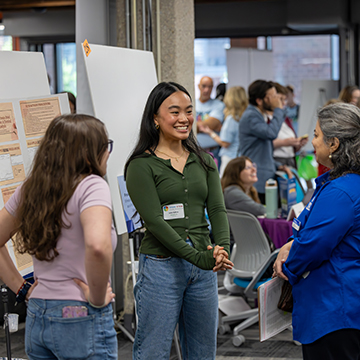 a group of attendees listen intently to a student present her research at the STEM Poster Symposium