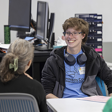 a smiling student sits at a table talking to two other individuals who are facing away from the viewer