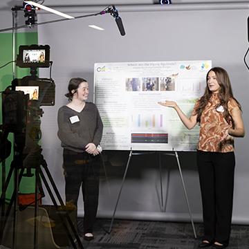 two students give their poster presentation in front of green screen while another student records them on a professional camera