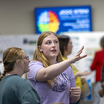 a student gestures at her poster while explaining her research to a symposium attendee