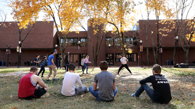 A group of students sitting on the ground watch other play Frisbee.