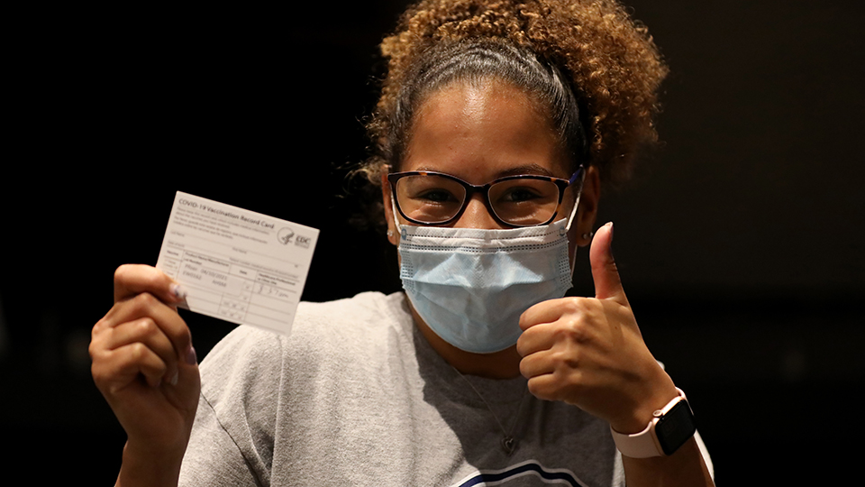 A woman holding a vaccination card and wearing a mask gives a thumbs up.