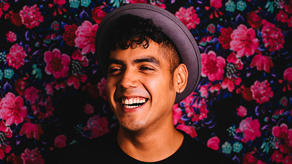 Portrait of Saul Flores wearing a hat in front of a hedge of pink flowers.